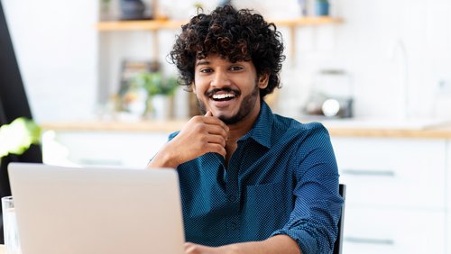 A young man with curly hair sits smiling in front of a laptop, resting his chin on his hand as he looks at the camera.