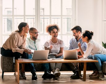 Sechs Personen sitzen um einen Tisch mit Laptops, lachen und diskutieren. Sie scheinen an einem kreativen oder geschäftlichen Projekt zu arbeiten. Der Raum ist hell und freundlich, mit großen Fenstern und Pflanzen im Hintergrund.