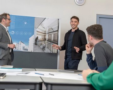 Ein Mann präsentiert ein Architekturprojekt vor einer Gruppe in einem Besprechungsraum. Er steht neben einem Plakat mit einem Gebäudeentwurf. Die Zuhörer, darunter ein Mann und eine Frau, zeigen Interesse an der Präsentation.