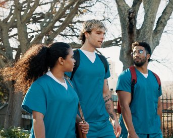 Three people in blue scrubs are walking together, surrounded by trees and a fence.