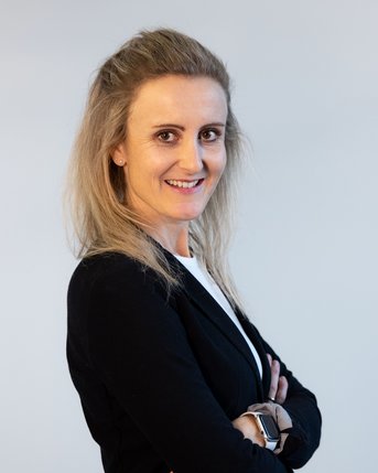 Eine Frau mit blonden Haaren in einem schwarzen Blazer steht mit verschränkten Armen und lächelt leicht, vor einem neutralen Hintergrund.