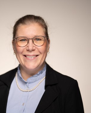 A woman with glasses and a pearl necklace is wearing a black blazer and a light blue shirt. She smiles warmly at the camera.