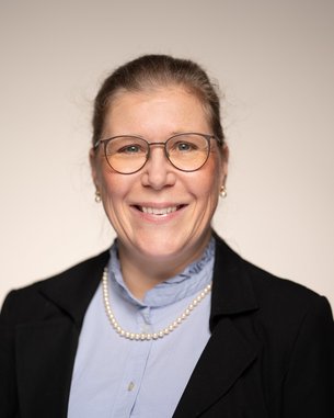 Eine lächelnde Frau mit Brille, blauer Bluse und schwarzem Blazer, steht vor einem neutralen Hintergrund.