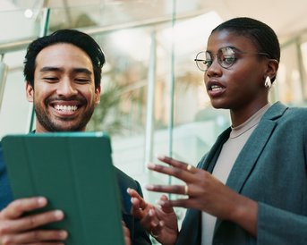 Ein Mann und eine Frau stehen in einem modernen Raum und diskutieren, während sie ein Tablet betrachten. Beide wirken engagiert.