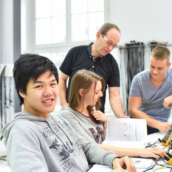 A group of four people in a lab working on technical equipment. A young man smiles at the camera while the others focus intently on their tasks.