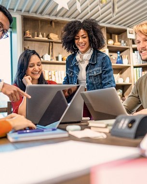 Eine Gruppe von fünf Personen sitzt an einem Tisch und schaut auf Laptops. Sie scheinen an einem Projekt zu arbeiten und diskutieren miteinander. Die Atmosphäre wirkt kollaborativ und freundlich.