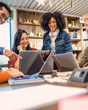 Eine Gruppe von fünf Personen sitzt an einem Tisch und schaut auf Laptops. Sie scheinen an einem Projekt zu arbeiten und diskutieren miteinander. Die Atmosphäre wirkt kollaborativ und freundlich.