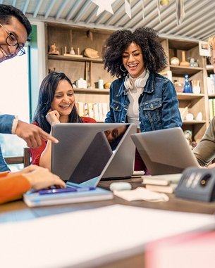 Eine Gruppe von fünf Personen sitzt an einem Tisch und schaut auf Laptops. Sie scheinen an einem Projekt zu arbeiten und diskutieren miteinander. Die Atmosphäre wirkt kollaborativ und freundlich.