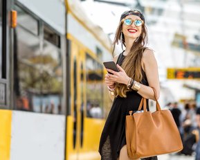Junge Frau in schwarzem Kleid und Sonnenbrille, hält ein Smartphone und eine große Handtasche, steht an einer Straßenbahnhaltestelle.