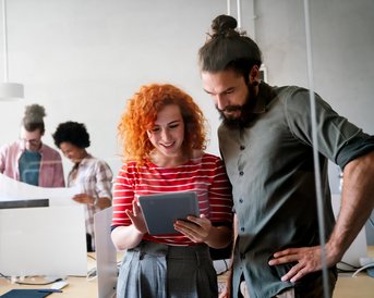 Two people are looking at a tablet together, while in the background, others are working on a project.
