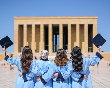 Vier Absolventinnen in blauen Roben und Hüten stehen mit dem Rücken zur Kamera vor einem monumentalen Gebäude.