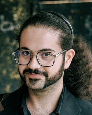 Mann mit lockigem Haar und Brille, trägt einen schwarzen Blazer. Er schaut direkt in die Kamera und hat einen gepflegten Bart.