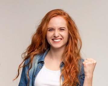 Eine lächelnde Frau mit roten Haaren zeigt eine Siegerpose, während sie in einem blauen Hemd und einem weißen T-Shirt gekleidet ist.