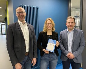 Drei Personen stehen in einem modernen Büro. Eine Person hält ein Buch in der Hand, während die anderen lächeln und in die Kamera schauen.