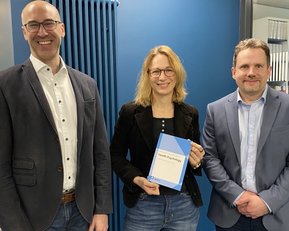 Three people are standing in a modern office. One person is holding a book, while the others are smiling and looking at the camera.