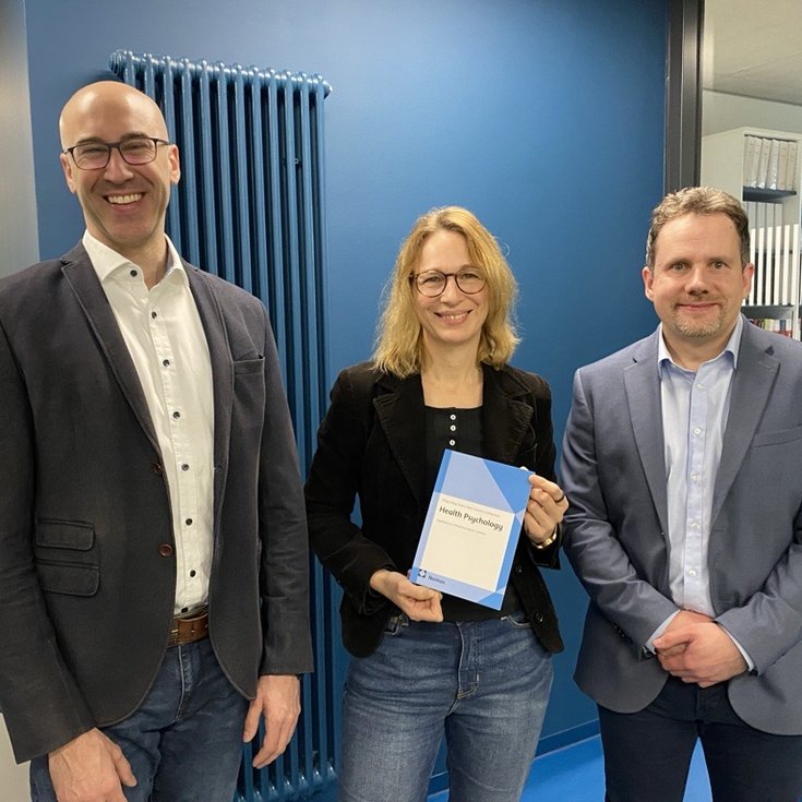 Three people are standing in a modern office. One person is holding a book, while the others are smiling and looking at the camera.