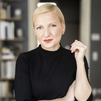 Eine Frau mit blonden Haaren und rotem Lippenstift trägt ein schwarzes Oberteil und posiert lächelnd in einem modernen Raum.