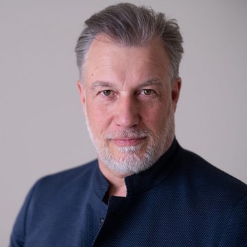 Ein Mann mit grauen Haaren und Bart trägt eine dunkle Jacke und schaut direkt in die Kamera. Der Hintergrund ist neutral.