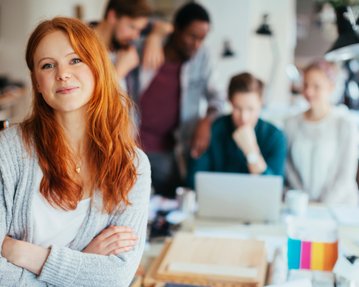 Eine lächelnde Frau mit roten Haaren steht im Vordergrund, während mehrere Personen im Hintergrund an einem Tisch arbeiten.