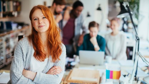 Eine lächelnde Frau mit roten Haaren steht im Vordergrund, während mehrere Personen im Hintergrund an einem Tisch arbeiten.