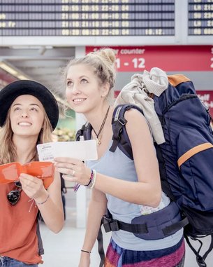 Zwei junge Frauen stehen in einem Flughafen und schauen auf eine Anzeigetafel. Eine Frau zeigt mit dem Finger nach oben, während die andere ein Ticket in der Hand hält. Beide tragen Rucksäcke und lächeln.