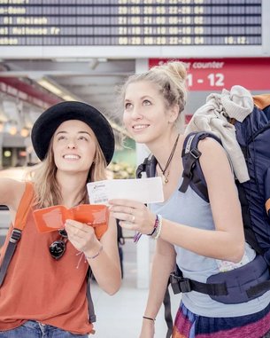 Zwei junge Frauen stehen in einem Flughafen und schauen auf eine Anzeigetafel. Eine Frau zeigt mit dem Finger nach oben, während die andere ein Ticket in der Hand hält. Beide tragen Rucksäcke und lächeln.