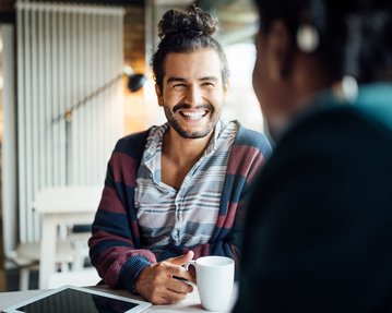 Ein Mann mit lockigem Haar und einem gestreiften Pullover sitzt an einem Tisch, lächelt und hält eine Tasse in der Hand.