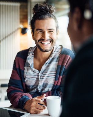 Ein Mann mit lockigem Haar und einem gestreiften Pullover sitzt an einem Tisch, lächelt und hält eine Tasse in der Hand.