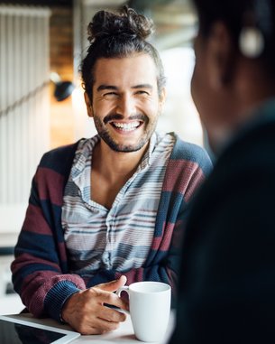 Ein Mann mit lockigem Haar und einem gestreiften Pullover sitzt an einem Tisch, lächelt und hält eine Tasse in der Hand.
