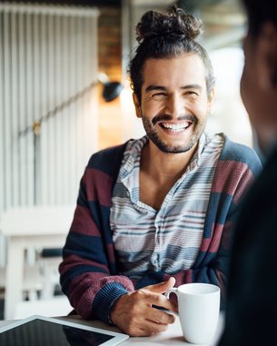 Ein Mann mit lockigem Haar und einem gestreiften Pullover sitzt an einem Tisch, lächelt und hält eine Tasse in der Hand.
