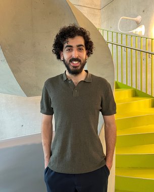 Ein Mann mit lockigem Haar steht lächelnd vor einer modernen Treppe mit gelben Stufen und einer grauen Wand im Hintergrund.