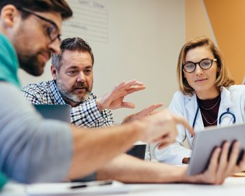 Eine Gruppe von Fachleuten diskutiert intensiv über Informationen auf einem Tablet während eines Meetings. Sie scheinen in einem medizinischen oder geschäftlichen Kontext zu arbeiten.