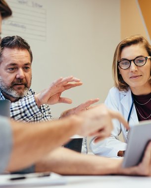 A group of professionals is intensely discussing information on a tablet during a meeting. They appear to be working in a medical or business context.