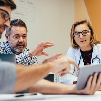 A group of professionals is intensely discussing information on a tablet during a meeting. They appear to be working in a medical or business context.