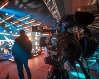 Eine Kamera steht im Vordergrund, während eine Person im Hintergrund auf eine beleuchtete Bühne blickt. Die Bühne ist mit bunten Lichtern und grafischen Elementen dekoriert, die auf eine bevorstehende Veranstaltung hinweisen.