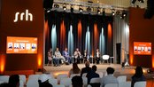 Eine Diskussionsrunde mit mehreren Teilnehmern auf einer Bühne, umgeben von Scheinwerfern und einem großen srh-Logo im Hintergrund. Zuschauer sitzen im Vordergrund.