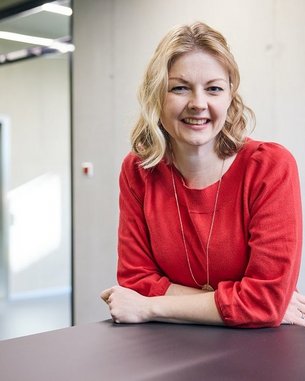 Eine lächelnde Frau mit blonden, lockigen Haaren trägt ein rotes Oberteil und lehnt an einem Tisch in einem modernen Raum.