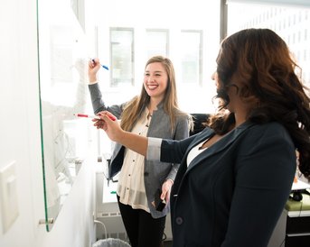Zwei Frauen stehen in einem Büro und schreiben an einem Glaswhiteboard. Eine Frau hält einen blauen Marker, während die andere einen roten Marker benutzt. Beide scheinen an einer Diskussion oder Präsentation beteiligt zu sein.