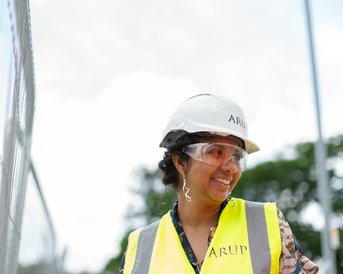 Eine lächelnde Frau in einem Sicherheitshelm und einer Warnweste steht an einer Baustelle. Im Hintergrund sind Bauzäune und Bäume sichtbar. Sie trägt eine Schutzbrille und wirkt freundlich und engagiert.