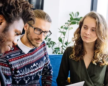 Drei Personen sitzen zusammen und diskutieren, während sie Laptops und Notizen verwenden. Eine Person zeigt auf ein Dokument.