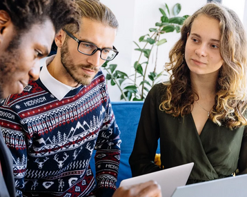 Drei Personen sitzen zusammen und diskutieren, während sie Laptops und Notizen verwenden. Eine Person zeigt auf ein Dokument.