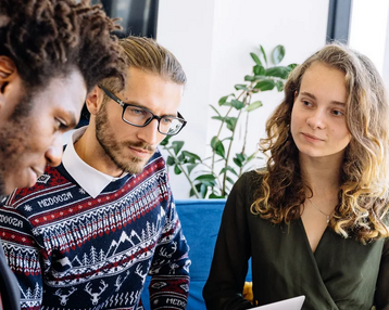 Drei Personen sitzen zusammen und diskutieren, während sie Laptops und Notizen verwenden. Eine Person zeigt auf ein Dokument.