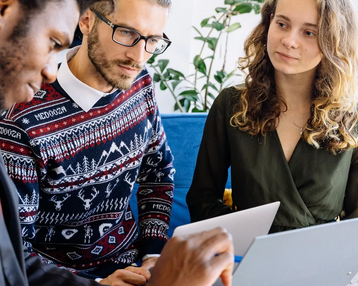 Drei Personen sitzen zusammen und diskutieren, während sie Laptops und Notizen verwenden. Eine Person zeigt auf ein Dokument.