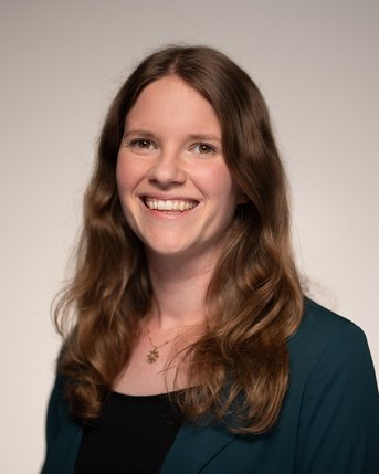 Eine lächelnde Frau mit langen, lockigen Haaren trägt ein schwarzes Oberteil und eine grüne Jacke. Der Hintergrund ist neutral.