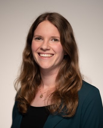 Eine lächelnde Frau mit langen, lockigen Haaren trägt ein schwarzes Oberteil und eine grüne Jacke. Der Hintergrund ist neutral.