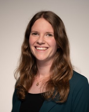 Eine lächelnde Frau mit langen, lockigen Haaren trägt ein schwarzes Oberteil und eine grüne Jacke. Der Hintergrund ist neutral.