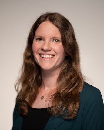 Eine lächelnde Frau mit langen, lockigen Haaren trägt ein schwarzes Oberteil und eine grüne Jacke. Der Hintergrund ist neutral.