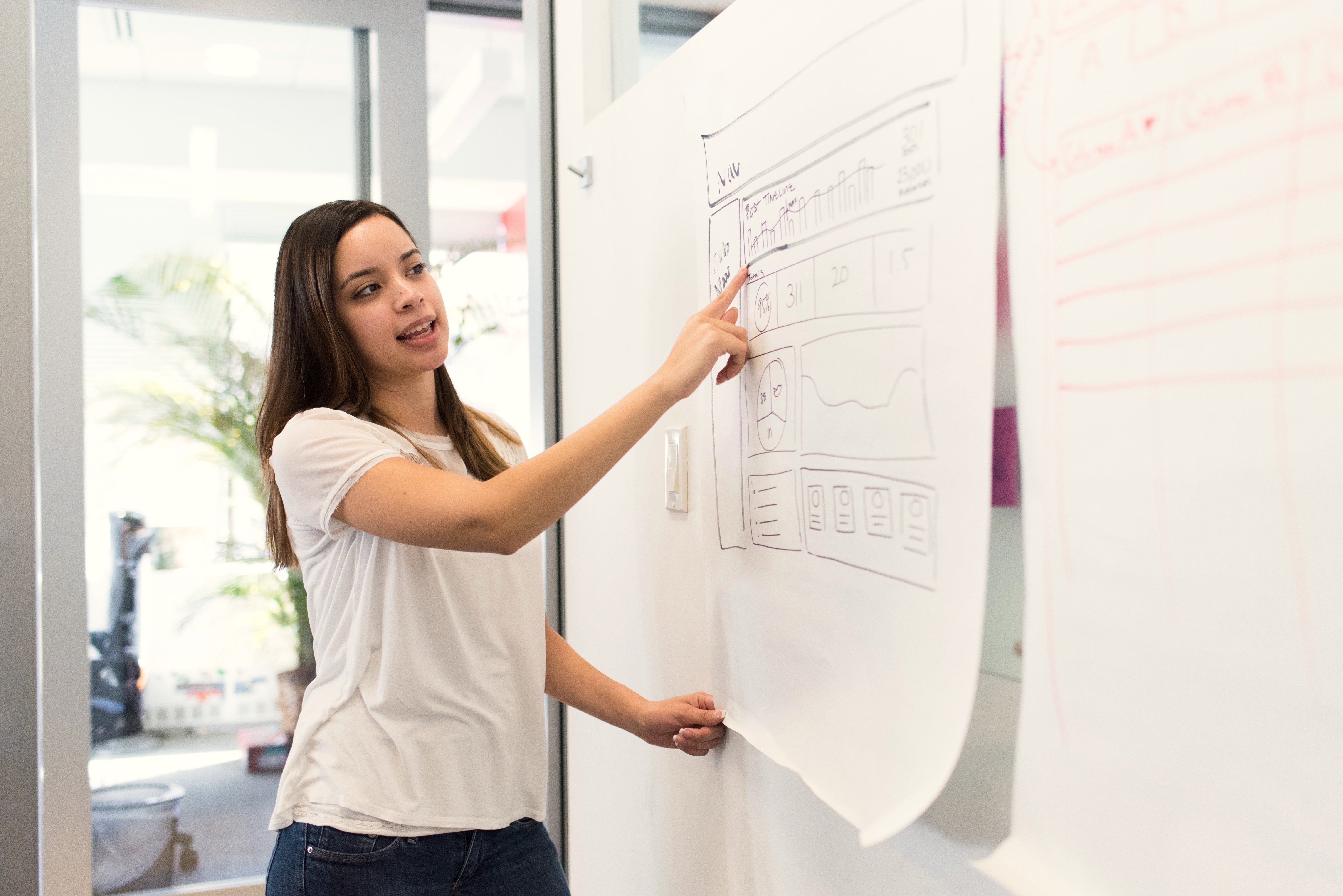 A woman points to a large sheet of paper displaying a graphical user interface or a diagram. She is wearing a simple, light-colored T-shirt and stands in a modern room with large windows.