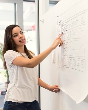 Eine Frau zeigt auf ein großes Papier, das eine grafische Benutzeroberfläche oder ein Diagramm darstellt. Sie trägt ein einfaches, helles T-Shirt und steht in einem modernen Raum mit großen Fenstern.