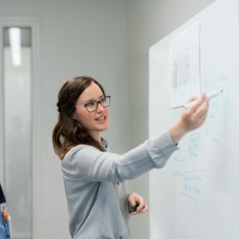 Eine Frau erklärt etwas an einem Whiteboard, während ein Mann aufmerksam zuhört. Beide sind in einem modernen Büro. Die Frau hat eine Brille und trägt einen grauen Pullover.